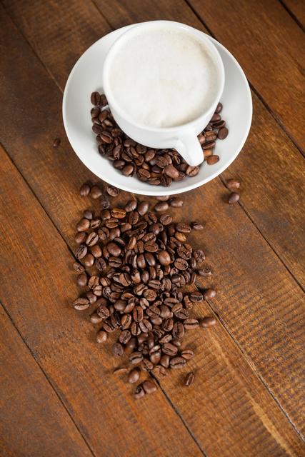 Cup of coffee with frothy top placed on white saucer filled with coffee beans. Additional coffee beans scattered on wooden table. Ideal for use in coffee shop promotions, cafe menus, breakfast advertisements, or articles about coffee culture and morning routines.