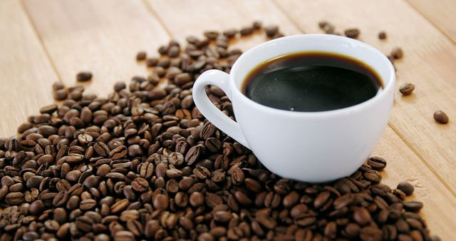 A white cup filled with coffee sits amidst a scattering of roasted coffee beans on a wooden surface. Coffee enthusiasts often appreciate the rich aroma and flavor that freshly brewed coffee offers.