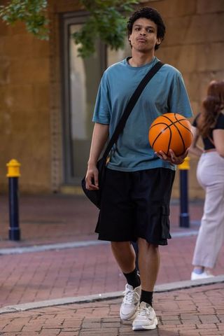 African american young man walking with basketball and crossbody bag on city sidewalk