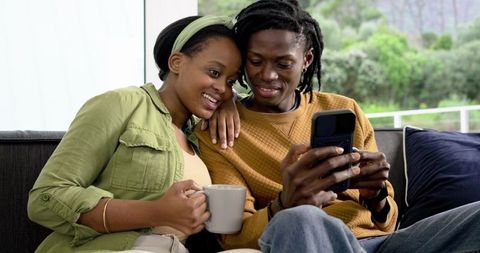African American Couple Relaxing on Sofa Sharing Coffee, Browsing Phone in Cozy Living Room