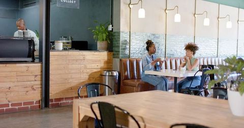 Two african american women chatting over coffee in modern rustic cafe booth with barista