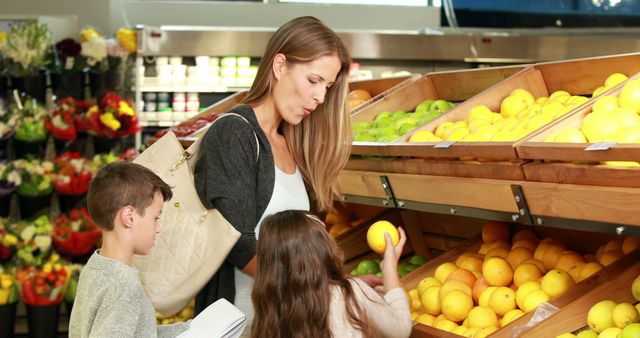 This image depicts a mother shopping in a grocery store with her children. She is examining citrus fruits, possibly oranges, while one child assists and the other writes on a notepad. The scene emphasizes family, healthy eating, and everyday activities. Useful for content featuring parenting, family life, grocery shopping habits, and healthy living.