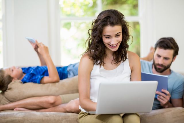 Young couple using digital devices in living room. Woman smiling while using laptop, man reading on tablet. Ideal for content on modern lifestyle, technology use, family relaxation, and home activities.