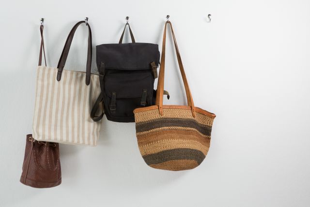 Assorted bags hanging on wall hooks, including a striped tote bag, a black backpack, a woven bag, and a brown leather bag. Ideal for use in articles or advertisements about fashion, home organization, interior design, or storage solutions.