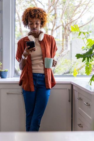 African american woman checking smartphone and sipping coffee at bright kitchen window