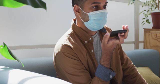 Man wearing a face mask sitting on a couch in an office, holding a smartphone near his mouth, and using the voice assistant feature. Useful for illustrations on pandemic lifestyle, technology use in office environments, remote work, and health safety measures.