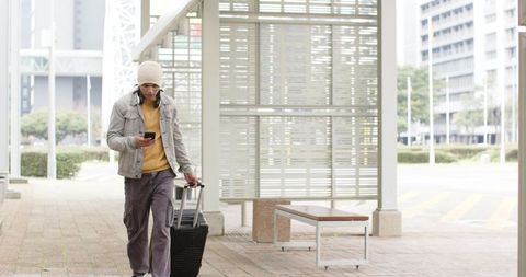 Young man walking through transit walkway pulling suitcase checking smartphone