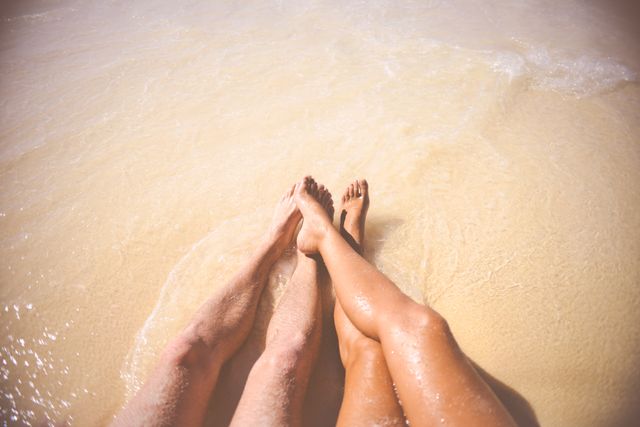 This images shows a couple's legs, tan and relaxed, on a sandy beach being gently touched by incoming waves. Perfect for travel brochures, romantic getaway promotions, summer vacation guides, beach resort advertising, and lifestyle blogs focusing on relationships or relaxation.