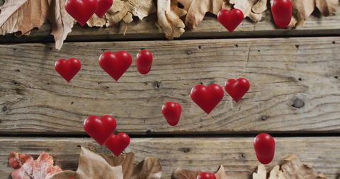 Floating glossy red hearts drifting over weathered wooden boards with autumn leaves closeup