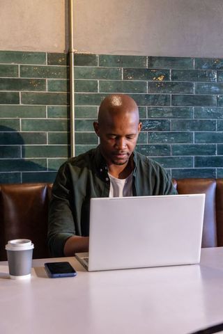 African american man working on laptop in urban cafe booth with coffee, smartphone