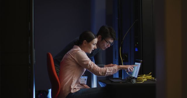 A man and woman are collaborating in a computer server room, analyzing data on a laptop. They appear focused and engaged in their work, with networking cables and servers visible around them. Ideal for illustrating themes of IT, technology, teamwork, and night shifts in professional environments.