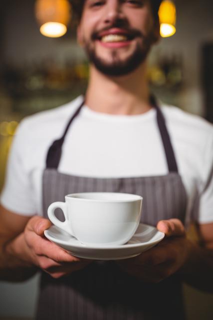Smiling barista holding a white porcelain coffee cup and saucer in a cafe. Ideal for use in articles and advertisements about coffee shops, customer service, barista training, and hospitality industry promotions.