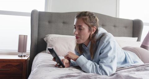 Young woman lying on bed holding smartphone in cozy pastel bedroom with tufted headboard soft light