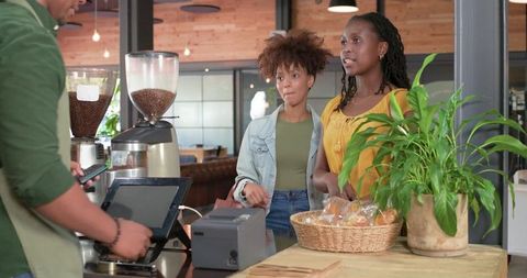 Two women ordering coffee and pastries at cozy modern cafe counter with friendly barista