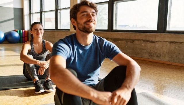Man and woman resting after workout in sunlit fitness studio with mats and large windows