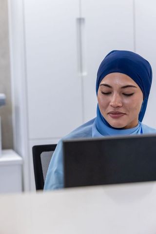 Female receptionist wearing hijab working at medical clinic front desk with laptop