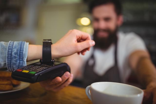 Woman paying for her purchase in a cafe using a smartwatch with NFC technology. Barista in background assisting with the transaction. Ideal for illustrating modern payment methods, digital wallets, and the convenience of contactless transactions in everyday settings.