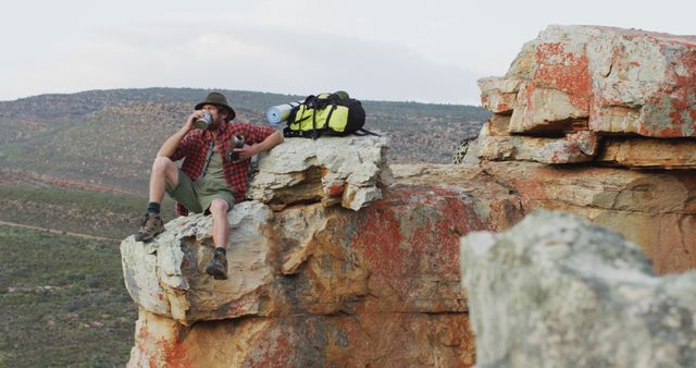 Caucasian male survivalist sitting on rocky mountain peak in wilderness, resting and drinking water - Download Free Stock Photos Pikwizard.com