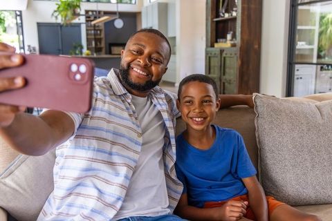 African american father and son taking selfie on couch, smiling, modern home lifestyle