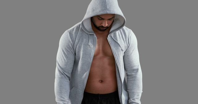 Athletic man in a gray hoodie stands with his head down, appearing focused and serious. His muscular physique is highlighted, suggesting he is preparing for a workout or training session. Useful in fitness and bodybuilding promotional materials, gym advertisements, exercise routines, or health blogs emphasizing physical strength and determination.