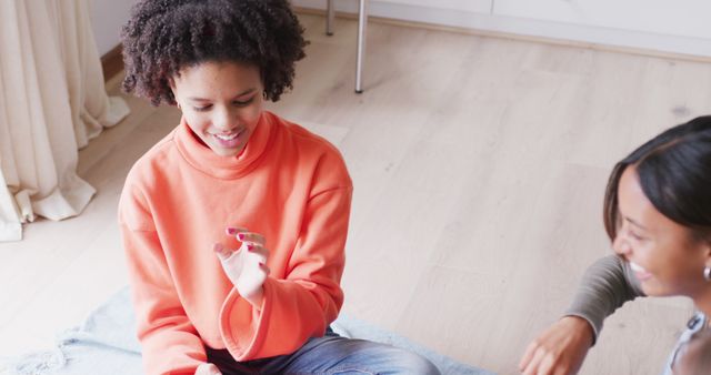 Two teen girls enjoying a casual and friendly conversation at home, sitting on a floor with happy expressions. Perfect for themes related to friendship, leisure activities, youth, communication services, and home life.