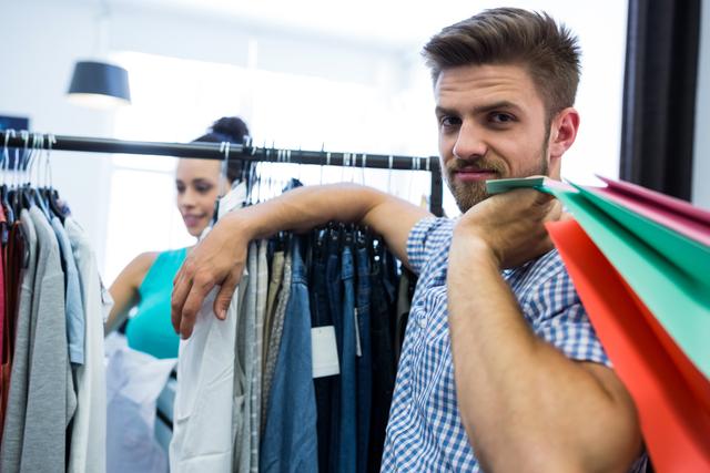 Man holding colorful shopping bags in a clothing store, smiling confidently. Ideal for use in retail advertisements, fashion promotions, lifestyle blogs, and articles about shopping experiences.