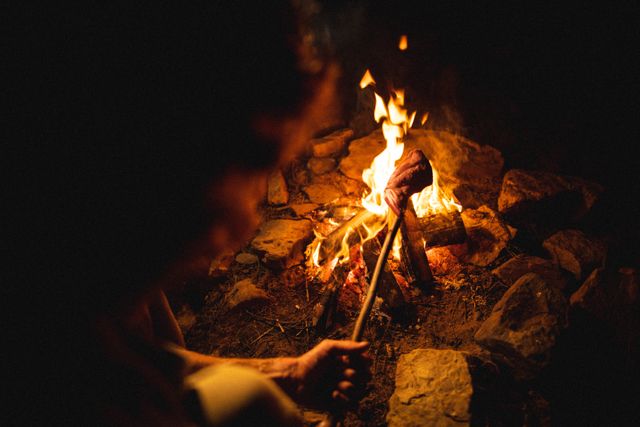 Young male survivalist roasting meat on a stick over a burning campfire in a forest at night. Ideal for content related to camping, survival skills, outdoor adventures, wilderness cooking, and weekend activities. Can be used in articles, blogs, and advertisements promoting outdoor experiences and survival techniques.