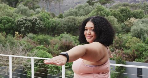 African American woman stretching on balcony overlooking lush greenery in pink tank