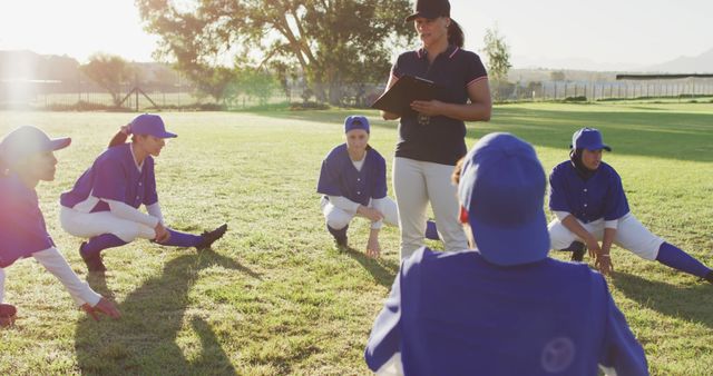 Diverse group of female baseball players with coach, warming up on pitch, squatting, stretching legs. female baseball team, sports training and game tactics.