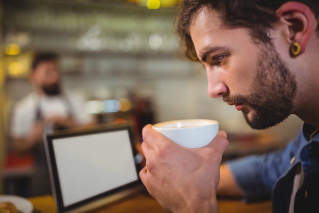 Young man drinking coffee while using a tablet in a cozy cafÃ©. Ideal for content related to leisure, technology, modern lifestyle, and coffee culture. Perfect for promoting cafes, digital services, and devices in a relaxed atmosphere.