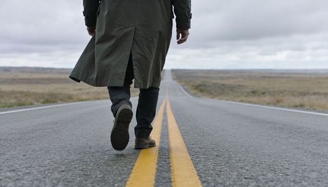 Man walking on empty open road toward distant horizon wearing trench coat and boots