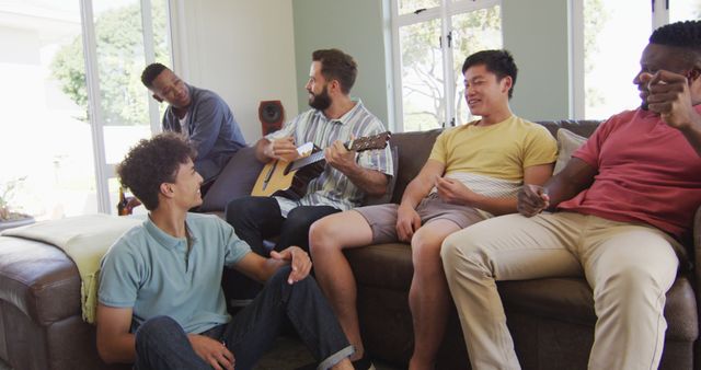Happy diverse male friends playing guitar and talking in living room. spending quality time at home together.