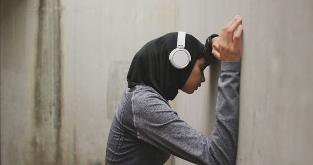Tired biracial woman in hijab wearing sports clothes and headphones leaning on wall in street. City living, fitness, music and modern urban lifestyle.