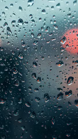 Vertical video capturing raindrops sliding on glass pane at dusk with red bokeh reflections