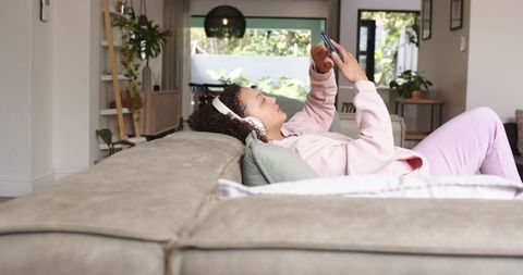 Young african american woman relaxing on sofa wearing headphones and scrolling phone at home