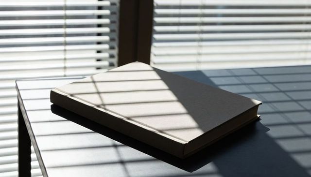 Sun casting blinds shadow across closed hardcover book on desk, minimalist workspace