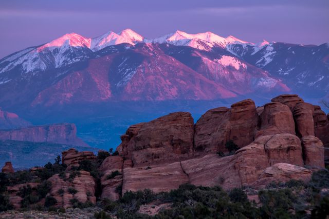 Beautiful capture of sunrise casting a pink glow on the La Sal Mountains with dry desert rocks in the foreground. This scene shows the stark, yet stunning, contrast between snow-covered peaks and rugged desert landscape. Perfect for illustrating natural beauty, serene moments, and adventurous travel destinations.