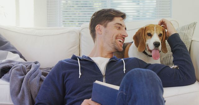 A young Caucasian man enjoys a relaxing moment on the couch with his beagle dog, with copy space. Their bond is evident as the man smiles at his pet, highlighting the companionship animals provide.
