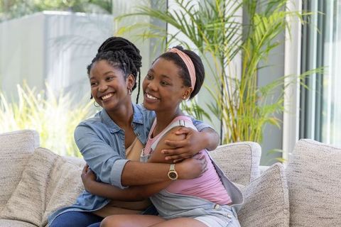 African mother and teenage daughter sharing warm hug on sunlit modern living room sofa