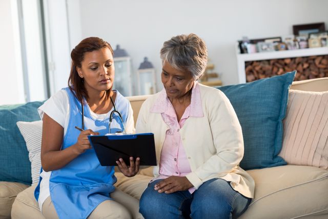 Doctor explaining medication on clipboard to senior woman in living room at home