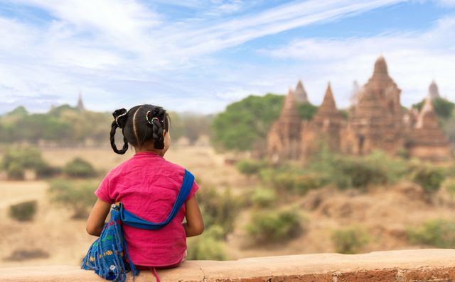 Young girl in red shirt with pigtails and traditional blue bag exploring ancient temples of Bagan, Myanmar. Great for travel and tourism content, cultural heritage site promotions, educational materials about Southeast Asia, and adventure travel marketing.