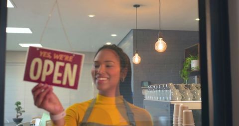 Smiling barista hanging open sign at cozy coffee shop window with warm pendant lighting