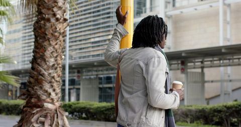 African American man waiting at plaza holding coffee leaning on yellow post by palm trees
