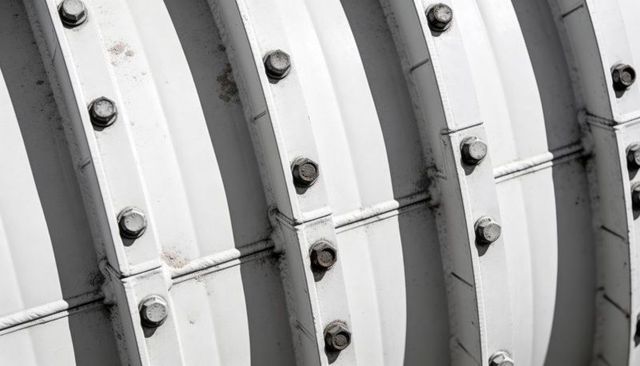 Curved industrial ductwork displaying bolted flanges, rusted hex bolts, geometric pattern