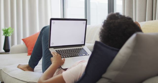 Person lounging on a sofa holding a laptop with a blank screen in a bright, comfortable living room. Useful for lifestyle or tech-related content, promoting home decor, illustrating work from home or casual browsing, and depicting contemporary living environments.
