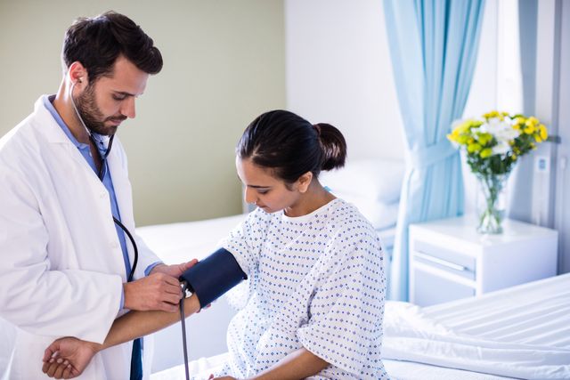 This image shows a male doctor checking the blood pressure of a female patient in a hospital room. The patient is wearing a hospital gown and sitting on a bed, while the doctor uses a stethoscope and blood pressure cuff. This image can be used for healthcare, medical, and hospital-related content, such as articles, brochures, and websites promoting patient care, medical services, and health checkups.