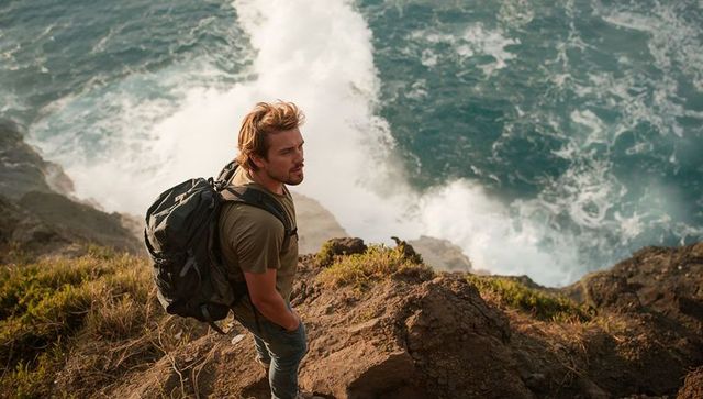 Solo male hiker standing on rocky coastal cliff gazing out over turquoise ocean waves