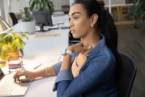 Young professional woman working at laptop touching neck in modern open-plan office