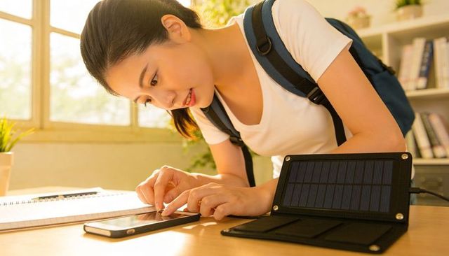 Student charging phone using portable solar powerbank on sunlit study desk with backpack