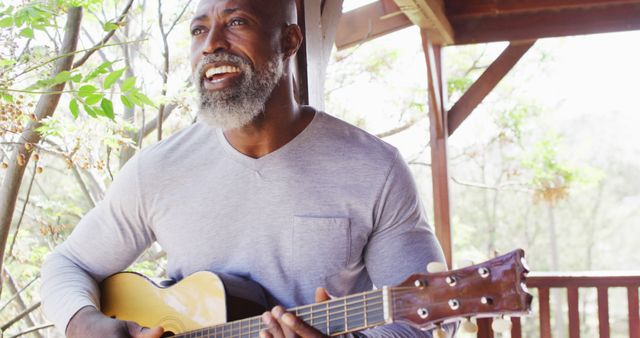 A mature man with a stylish beard is sitting on a porch playing an acoustic guitar. He appears to be smiling and enjoying the moment. Great for use in advertisements promoting relaxation, music lessons, retirement living, and moments of joy and leisure.